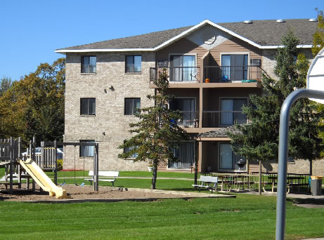 Multi-story residential building with beige stone facade, balconies, and sliding doors, overlooking a communal lawn area with playground and picnic tables under clear skies.